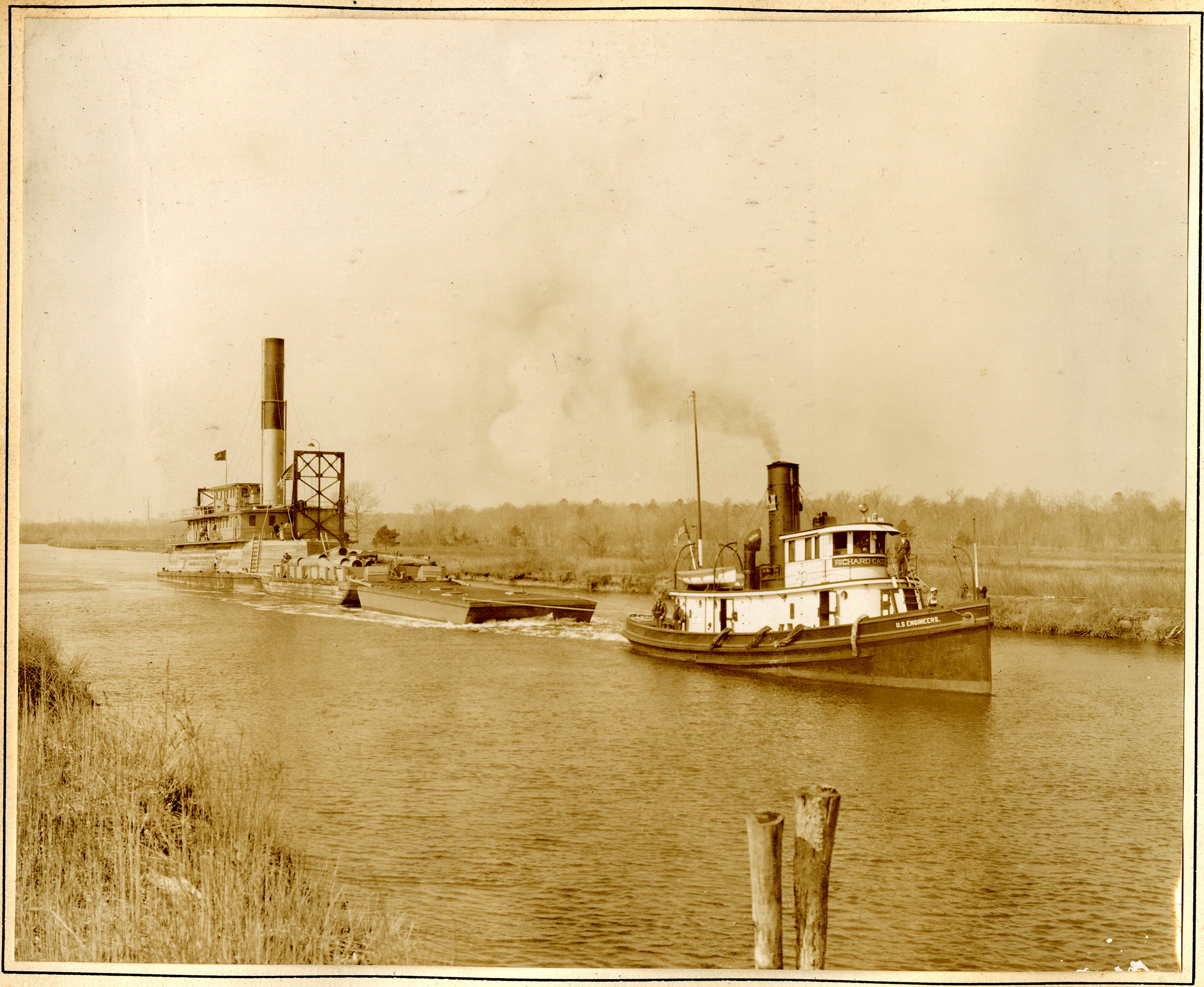 Tugboat is pulling a larger dredge behind it through a canal